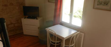 Una habitación pequeña con una mesa y sillas blancas, un televisor sobre un aparador y una ventana con cortina roja.