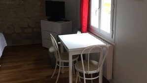 A small, well-lit room with a white table and chairs, a television on a white cabinet, and a window with a red curtain.