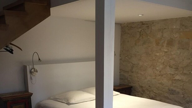 A bedroom with a wooden floor, a bed with white bedding, a stone wall, and a wooden nightstand with a lamp.