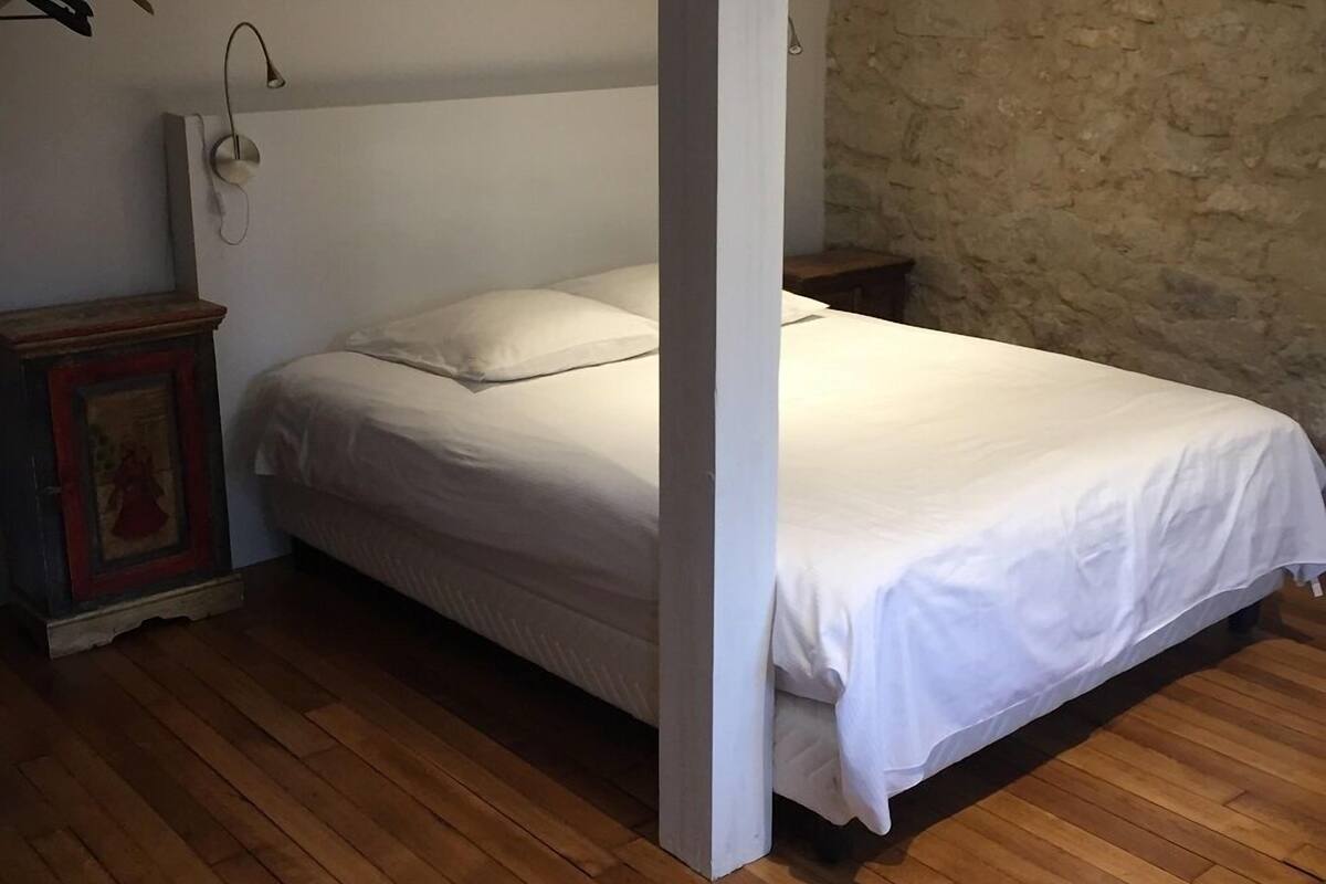 A bedroom with a wooden floor, a bed with white bedding, a stone wall, and a wooden nightstand with a lamp.