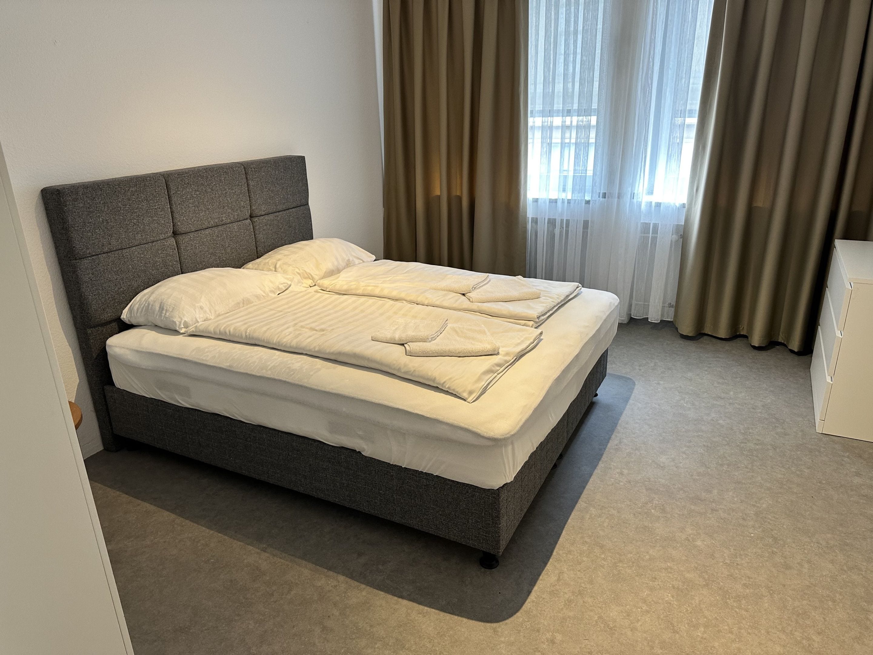 Ein Bett mit grauem Kopfteil, weißer Bettwäsche und Blick ins Freie durch transparente Vorhänge.