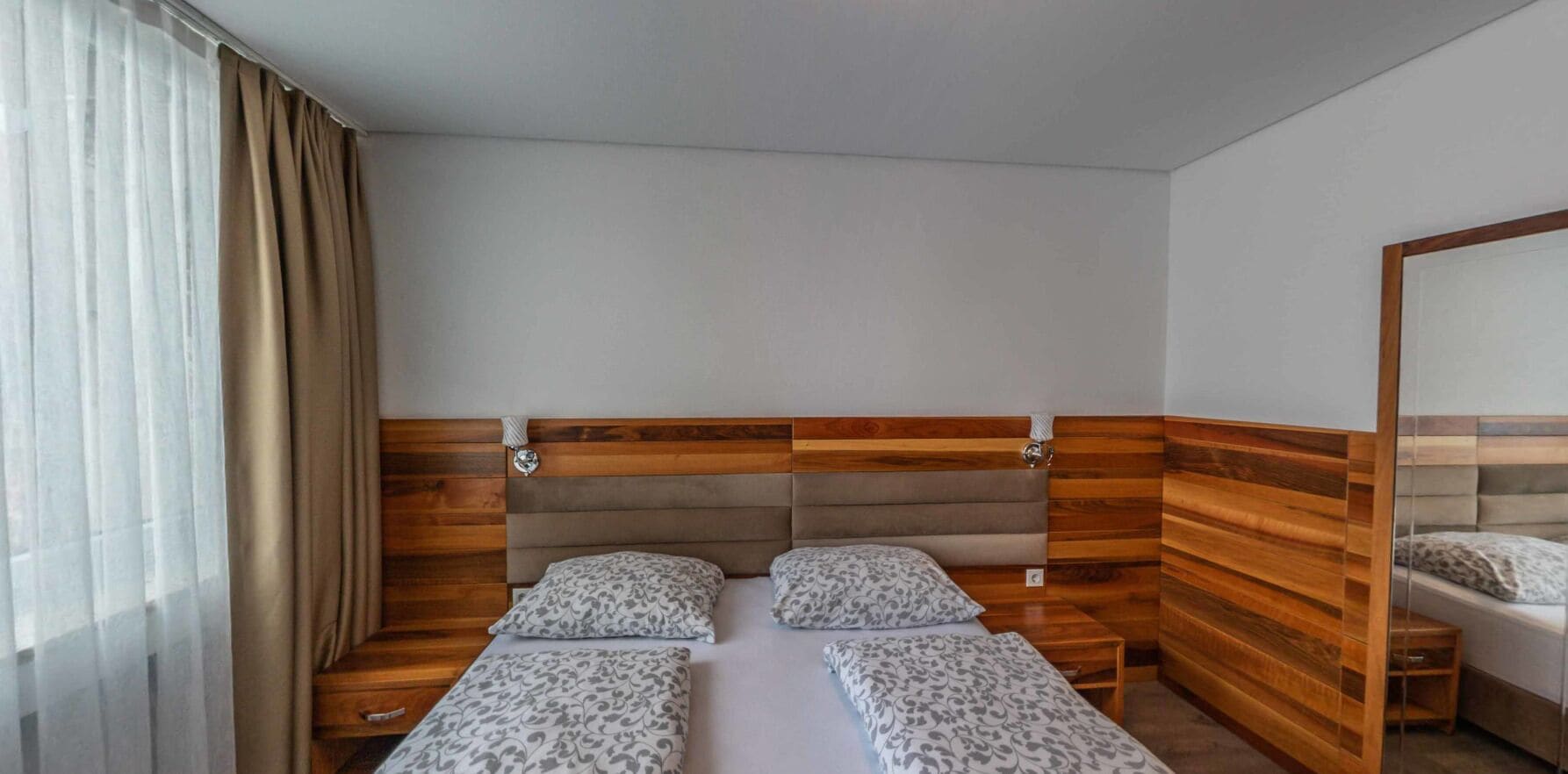 A hotel room with a wooden headboard, two beds with matching pillows, a bedside table, and a mirror.