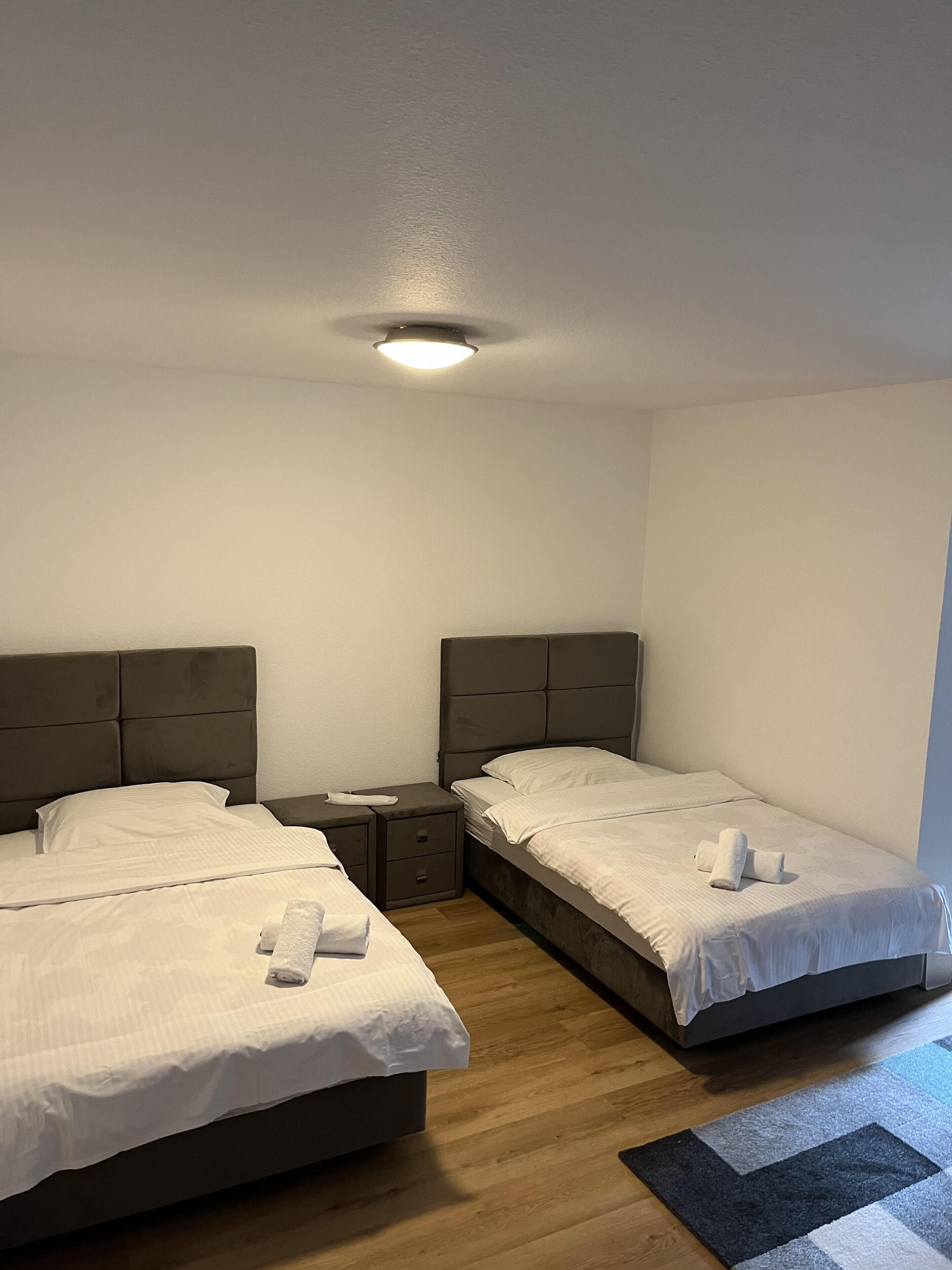 Habitación de hotel con dos camas, una mesita de noche, piso de madera y una lámpara en el techo.