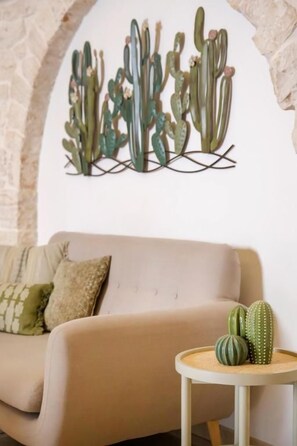 A living room with a beige sofa, a small round table with a cactus, and a wall-mounted cactus art piece. - Trulli Di Zia Vittoria (Alberobello)