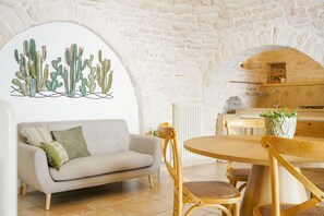 A cozy living room with a white sofa, wooden dining table, and chairs. There's a cactus wall art and a potted plant on the table. - Trulli Di Zia Vittoria (Alberobello)