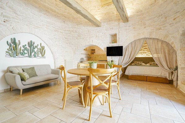 A cozy room with a stone wall, a wooden dining table with chairs, a sofa, a TV mounted on the wall, and a bed with a canopy. - Trulli Di Zia Vittoria (Alberobello)