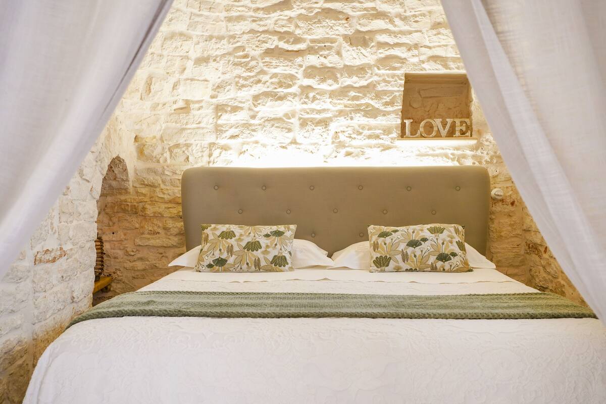 A bedroom with a stone wall, a bed with floral pillows, and a 'LOVE' sign above the bed.
