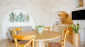 Een eetgedeelte met een ronde houten tafel, vier houten stoelen, wanddecoratie met een cactus, een potplant en een aan de muur bevestigde televisie.