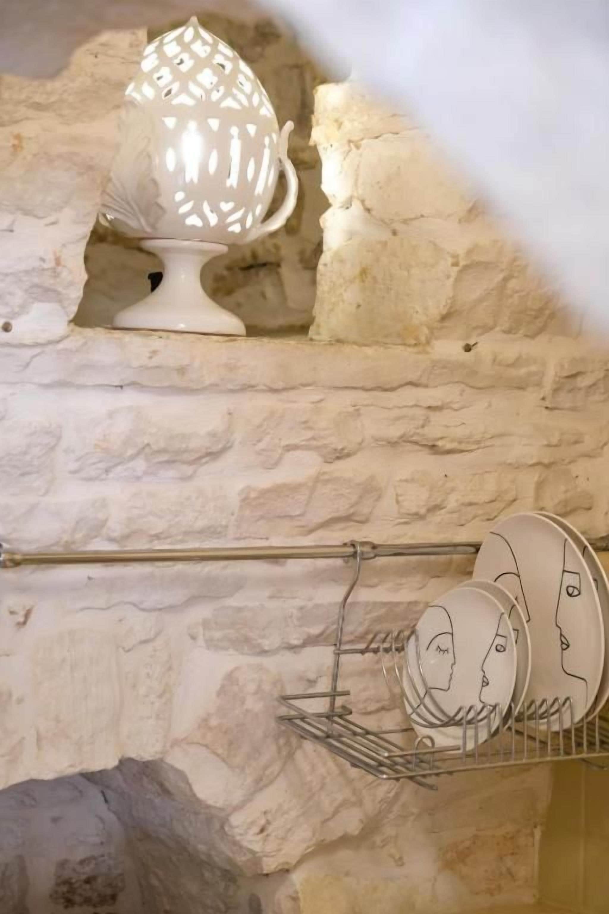 Una jarra de cerámica blanca sobre una estantería, un escurreplatos metálico y una pared de piedra.