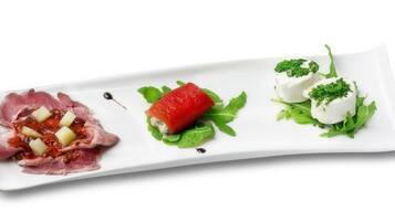 Plat blanc rectangular amb tres aliments: una làmina de carn amb cubs, una fragaria i dues culleradetes de crema blanca amb guarnició verda.