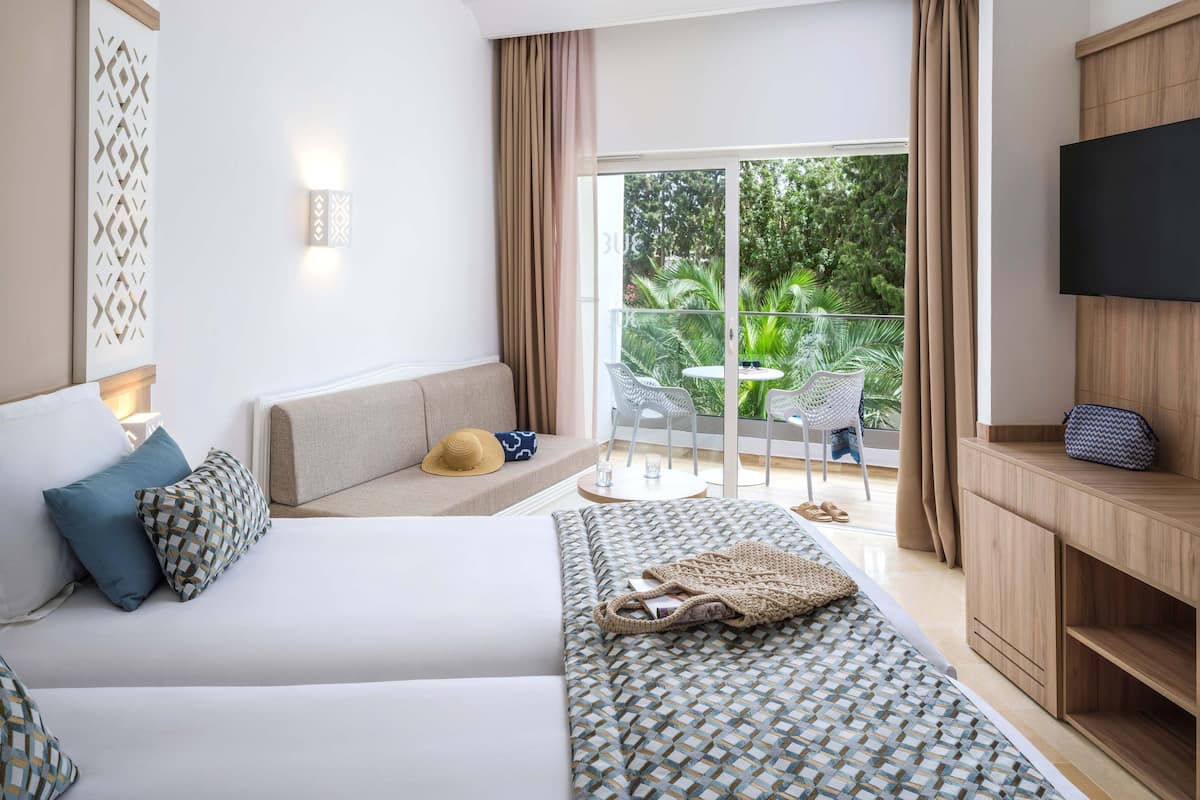 A hotel room with a bed, a sofa, a TV, a balcony with a table and chairs, and a view of greenery.