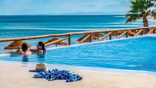 A poolside area with a wooden railing, a hat, and a towel on the sand.