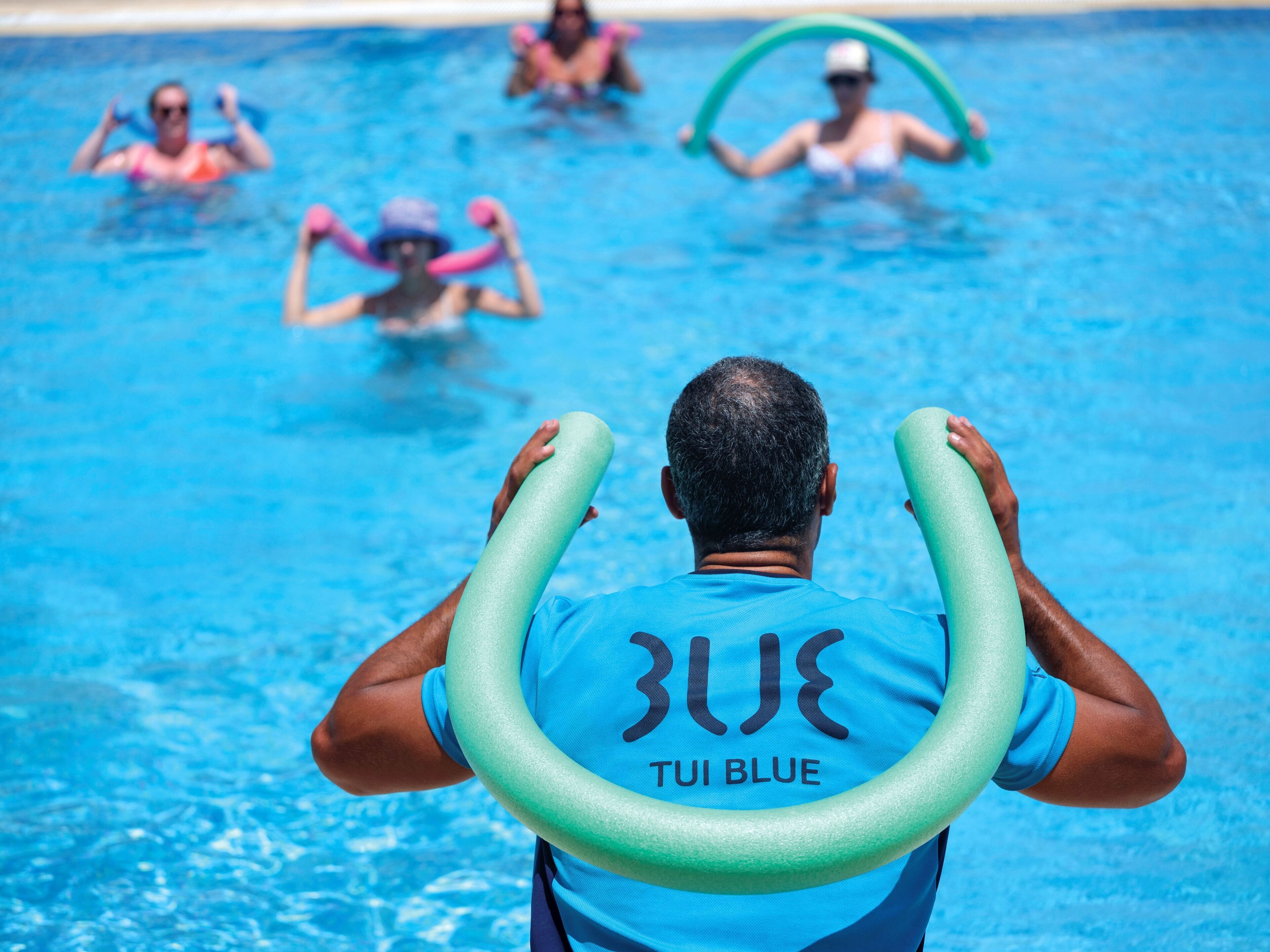 A man in a blue swim cap and shirt holding a green floatation device in a swimming pool with other people in the background.