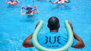 A man in a blue swim cap and shirt holding a green floatation device in a swimming pool with other people in the background.