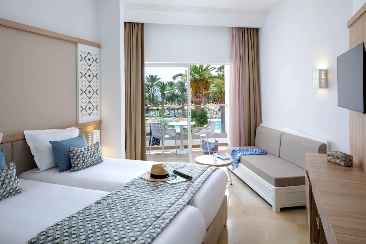 A hotel room with two beds, a sofa, a TV, and a balcony with a pool view.
