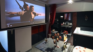 Una habitación de hotel moderna con una pantalla grande mostrando una película, una mesa de comedor con velas y una copa, y un elegante espacio de baño con tocador y espejo.