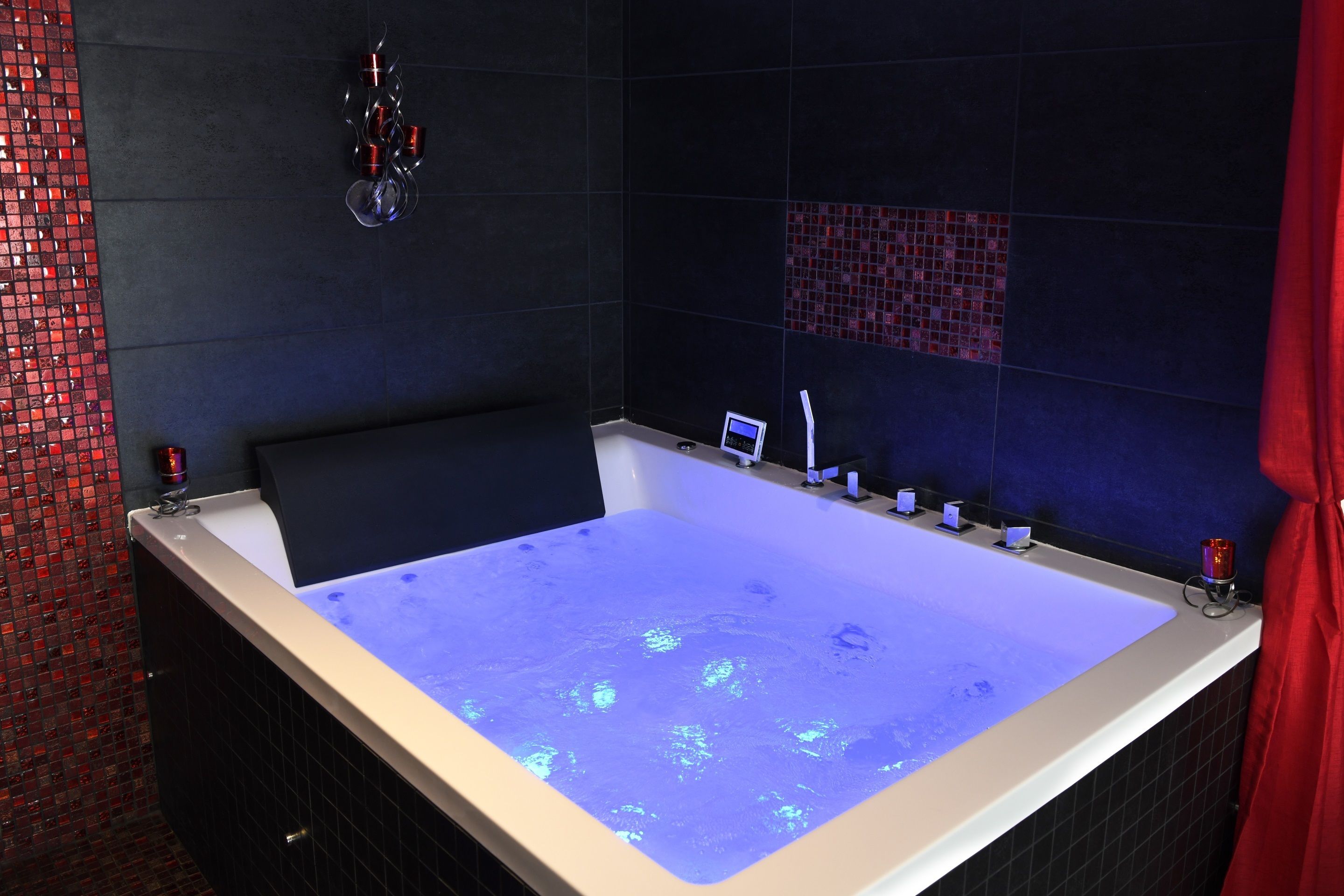 A large, rectangular bathtub with a digital display and a control panel, surrounded by dark tiles and a red curtain.
