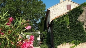 Una casa de piedra con camino de grava, rodeada de follaje y plantas en flor.