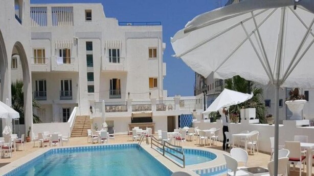 Un área de piscina con sillones blancos y sombrillas, una piscina azul cristalina y un edificio blanco con balcones al fondo.