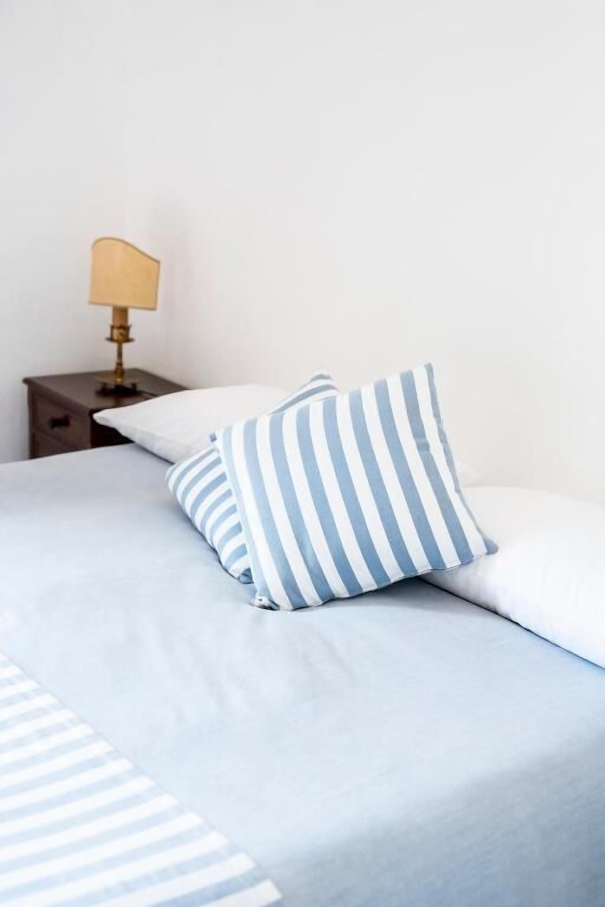 Un letto con un cuscino a righe e un cuscino semplice, un comodino con una lampada, e una parete neutra.