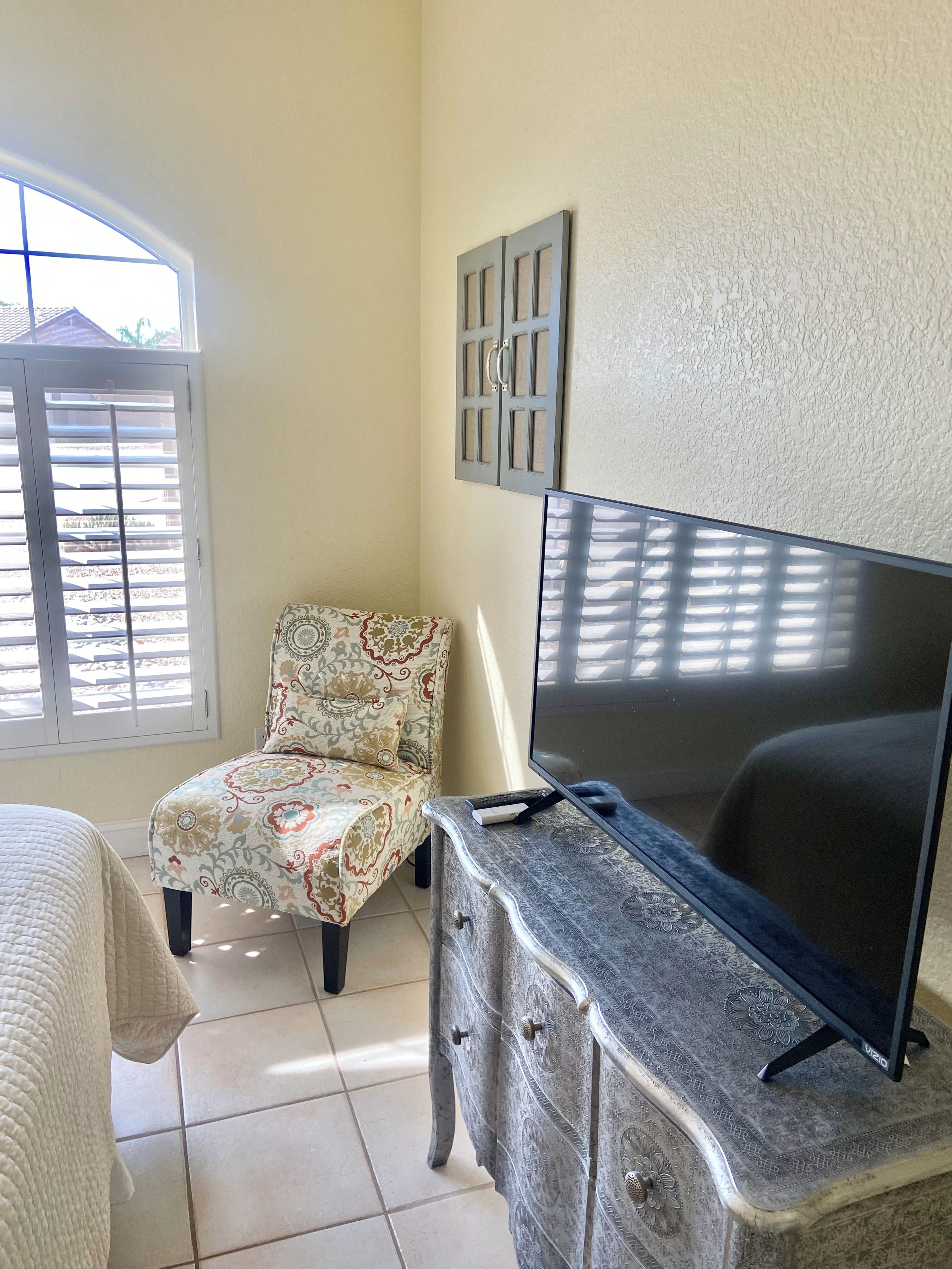 A bedroom with a bed, a chair, a dresser, a TV, and a window with blinds.