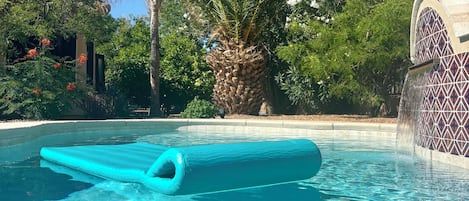 Une piscine aux eaux cristallines, avec un tapis flottant bleu, entourée d’une végétation luxuriante et d’un mur décoratif.