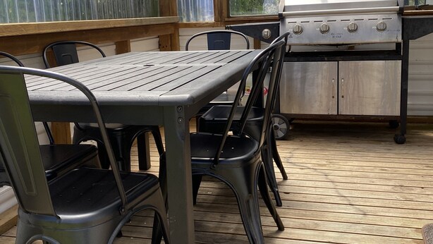 Eine Terrasse im Freien mit Holztisch und -stühlen, einem Grill und Blick auf Grün hinter den Fenstern.
