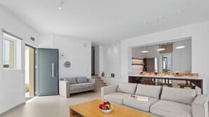 A modern living room with a sofa, coffee table, and a dining area with chairs and a kitchen counter.