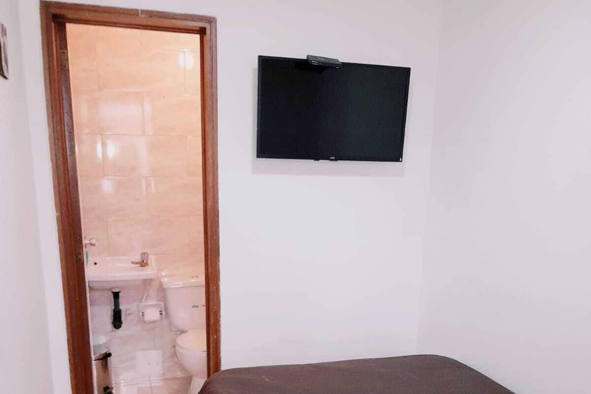A small, single-bed room with a wooden floor, a dark-colored bed, a blue pillow, a mounted TV, and a door leading to a bathroom with a toilet and sink.