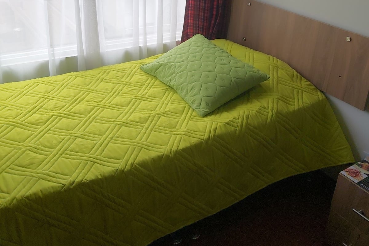 A single bed with a yellow quilt and a green pillow in a room with a wooden headboard and a window with sheer curtains.
