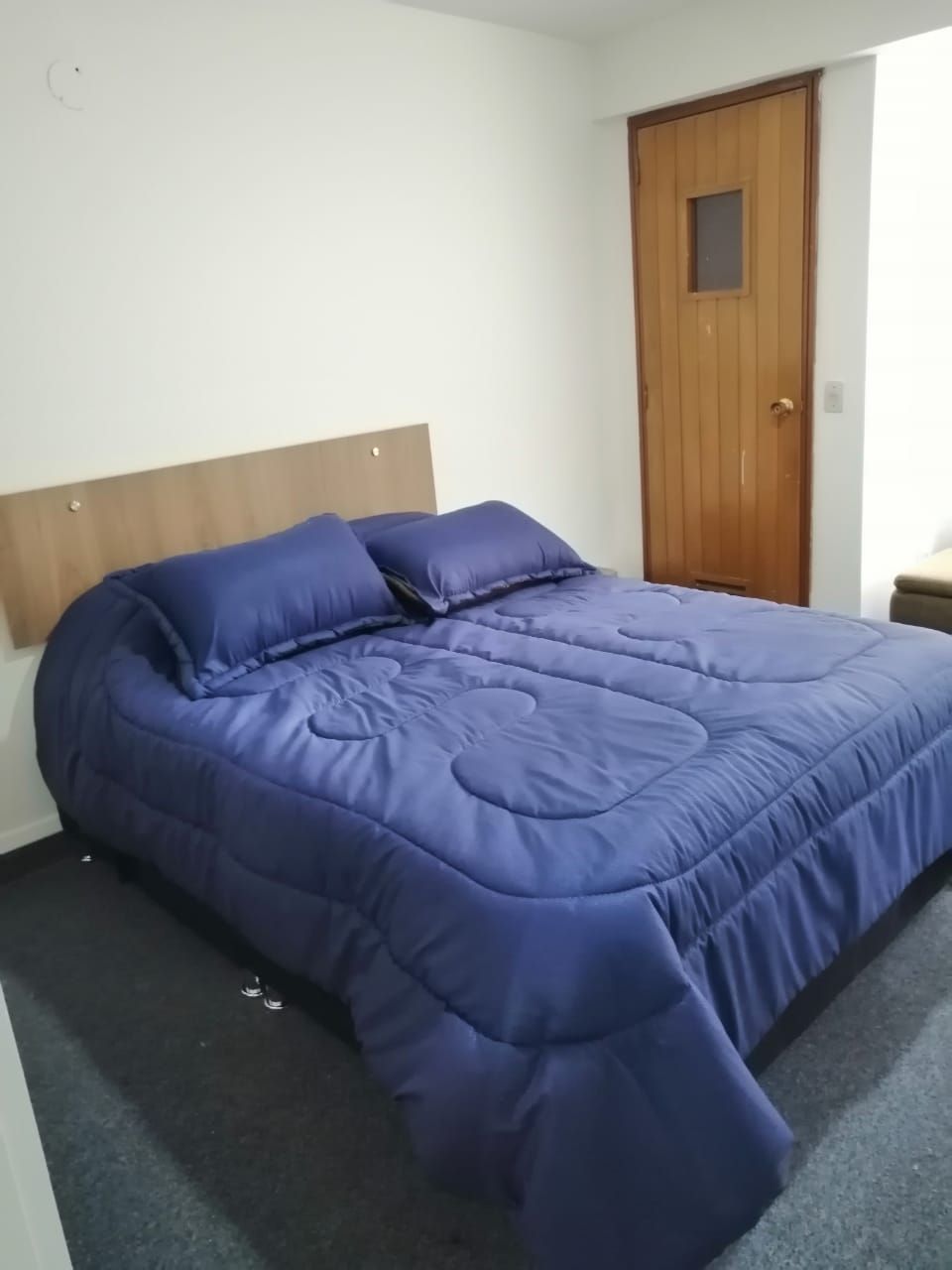 A single bed with a wooden headboard and a blue bedspread in a room with a wooden door and white walls.