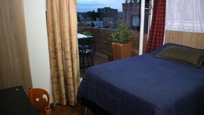 Un dormitorio con una cama grande, una mesita y una silla. Hay una ventana con cortinas y un balcón con vistas a la ciudad.