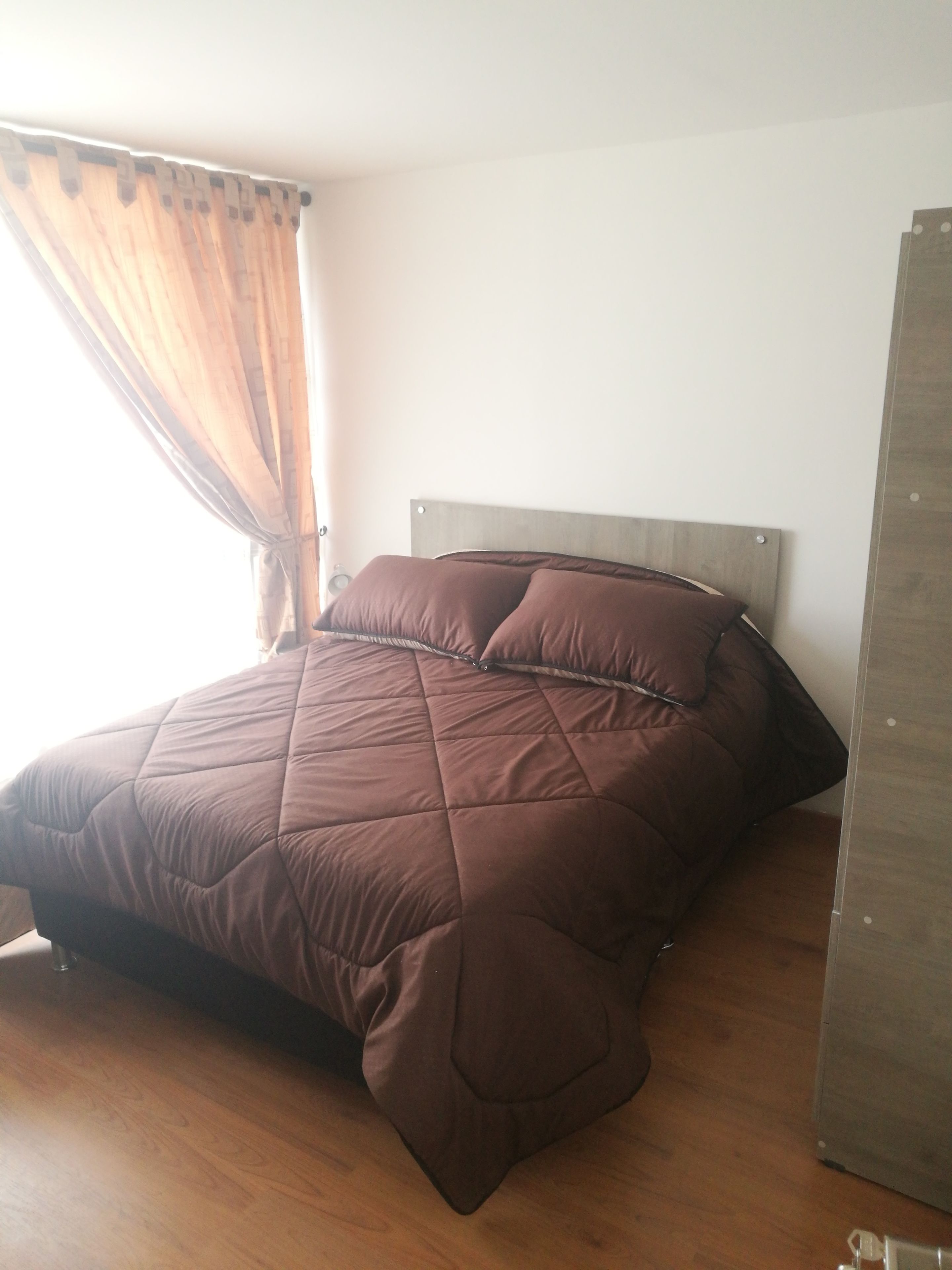 A bed with a brown quilt in a room with wooden flooring and a plain white wall.