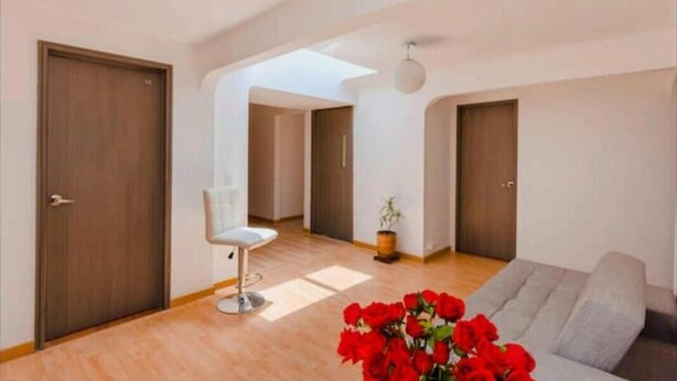 Amplia sala de estar con un sofá gris, una silla blanca y piso de madera. Hay dos puertas de madera y una pequeña planta en maceta.