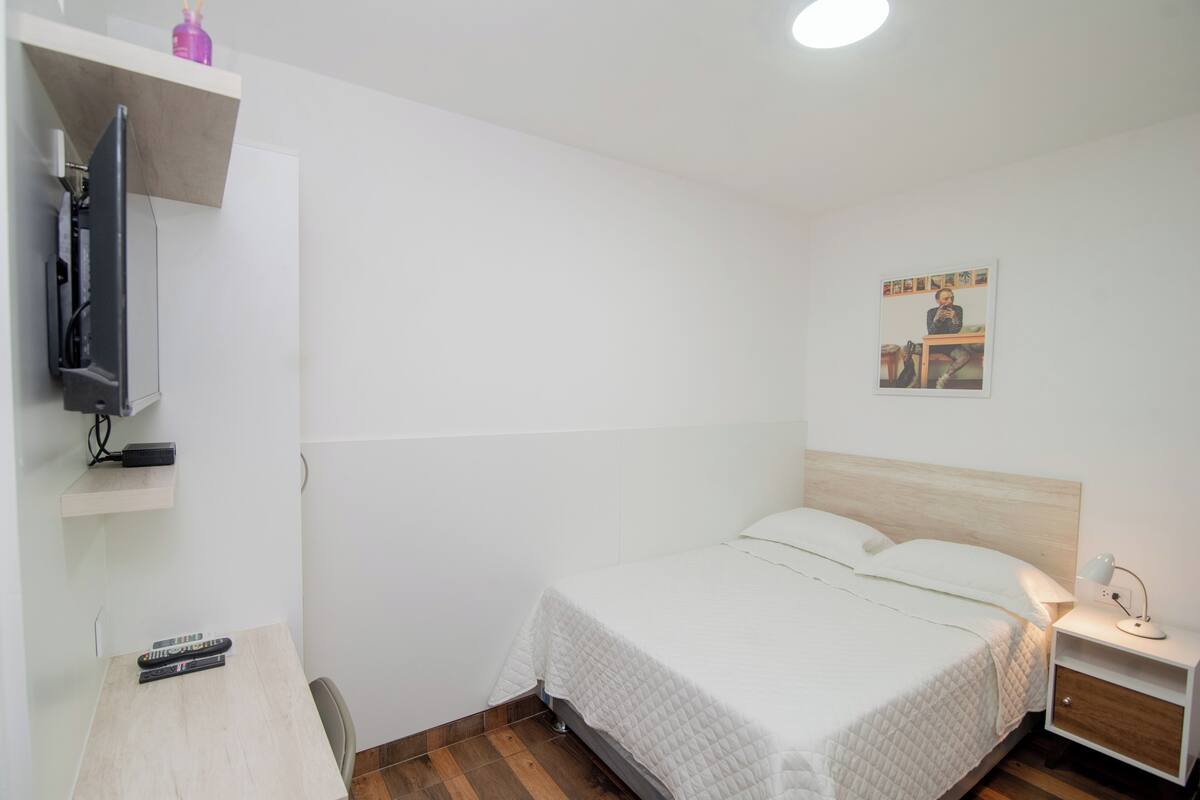 Quarto de hotel limpo e minimalista com uma cama de solteiro, uma escrivaninha pequena com cadeira, televisão na parede e mesa de cabeceira com abajur.
