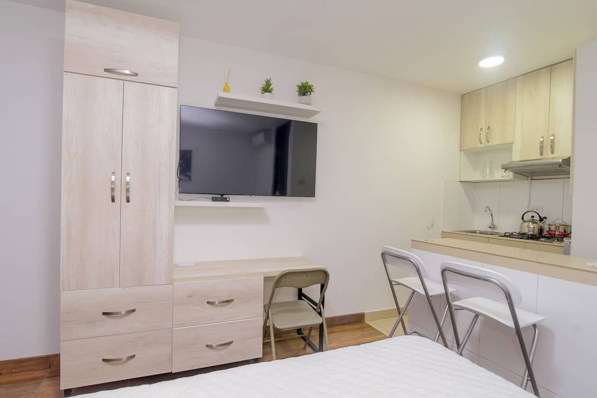 Quarto compacto com cama, escrivaninha e cadeira. Há uma TV fixada na parede acima da escrivaninha.