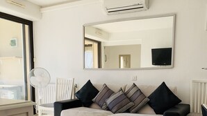 Una sala de estar con un sofá beige, una mesa de centro de vidrio, una mesita auxiliar blanca, un ventilador de pie y un equipo de aire acondicionado.
