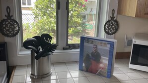 Uma cozinha com janela, um recipiente de metal com utensílios, um micro-ondas e um livro sobre o balcão.