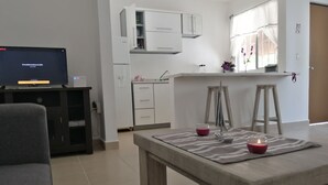 A modern living room with a flat-screen TV, a sofa, a dining table with a placemat, and a kitchen area with cabinets and a window. - Violet House - Riviera Maya (Puerto Morelos)