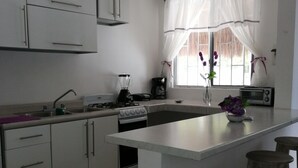 A modern kitchen with white cabinets, a central island, a toaster, and a window with curtains. - Violet House - Riviera Maya (Puerto Morelos)