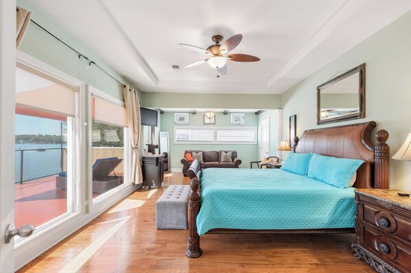 A bedroom with a bed, a ceiling fan, a large window, a balcony, and a desk with a chair.
