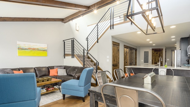 A modern living room with a sectional sofa, a blue armchair, and a dining table with chairs. A staircase with a metal railing leads to an upper level. - Hot Tub /Pool! Fireplace! Walk to Slopes! (Steamboat Springs)
