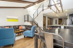 A modern living room with a sectional sofa, a blue armchair, and a dining table with chairs. A staircase with a metal railing leads to an upper level. - Hot Tub /Pool! Fireplace! Walk to Slopes! (Steamboat Springs)