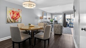 Un salon moderne avec une table à manger ronde, des chaises recouvertes d’un tissu gris, un canapé gris et un pouf à motifs. Un grand tableau abstrait orne le mur, et un luminaire est suspendu au plafond.