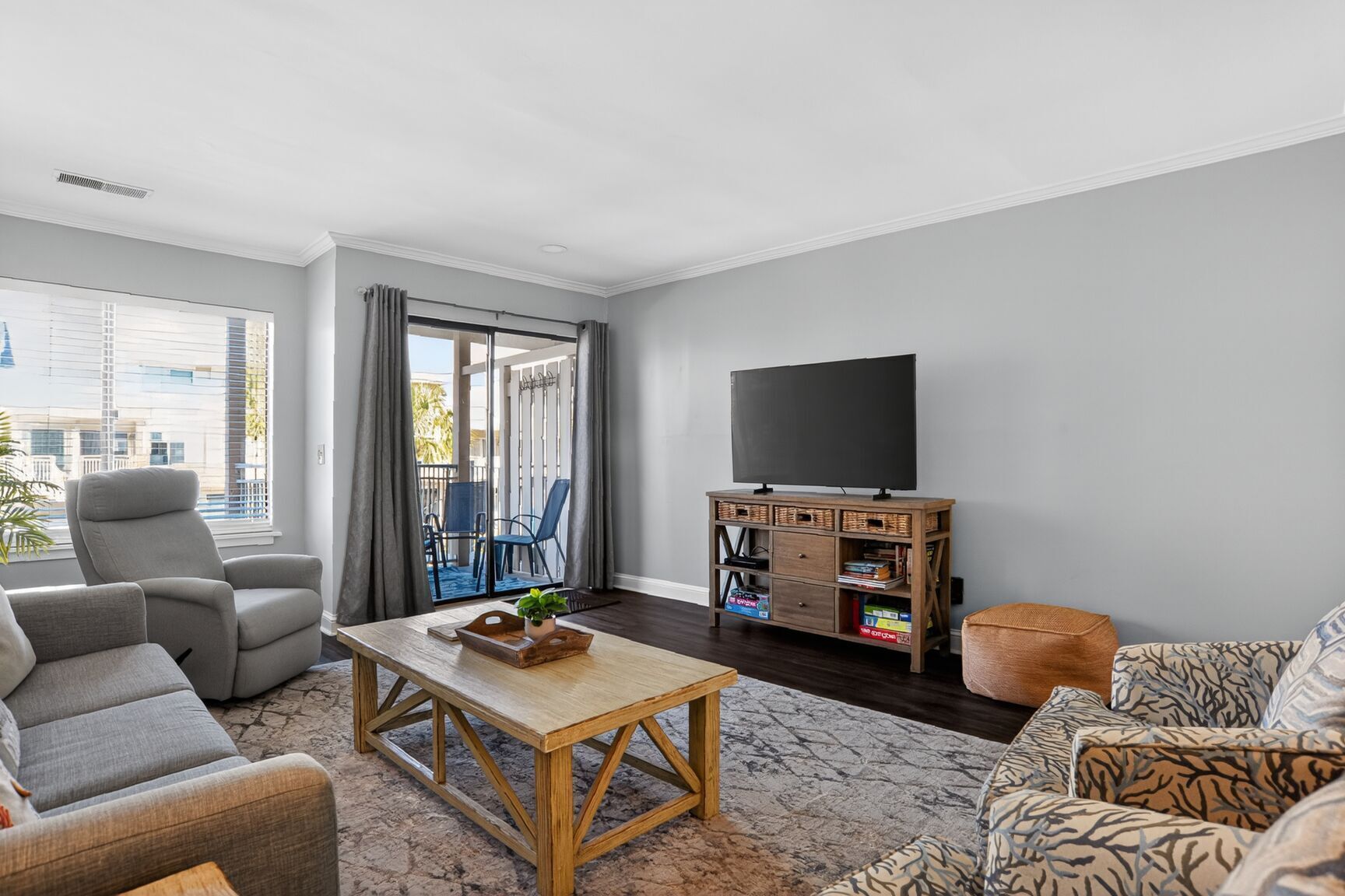 Una sala de estar con sofá, sillón y una mesa de centro. Hay un televisor sobre un mueble de madera y un balcón con vistas a edificios.