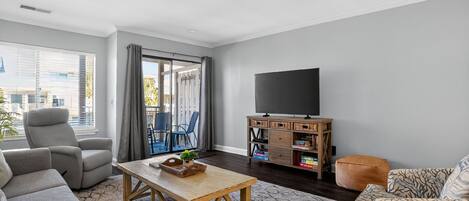 Una sala de estar con sofá, sillón y una mesa de centro. Hay un televisor sobre un mueble de madera y un balcón con vistas a edificios.