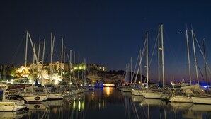 Un puerto deportivo al anochecer con varios veleros amarrados, un edificio destacado con luces en el fondo, y agua tranquila que refleja las luces.