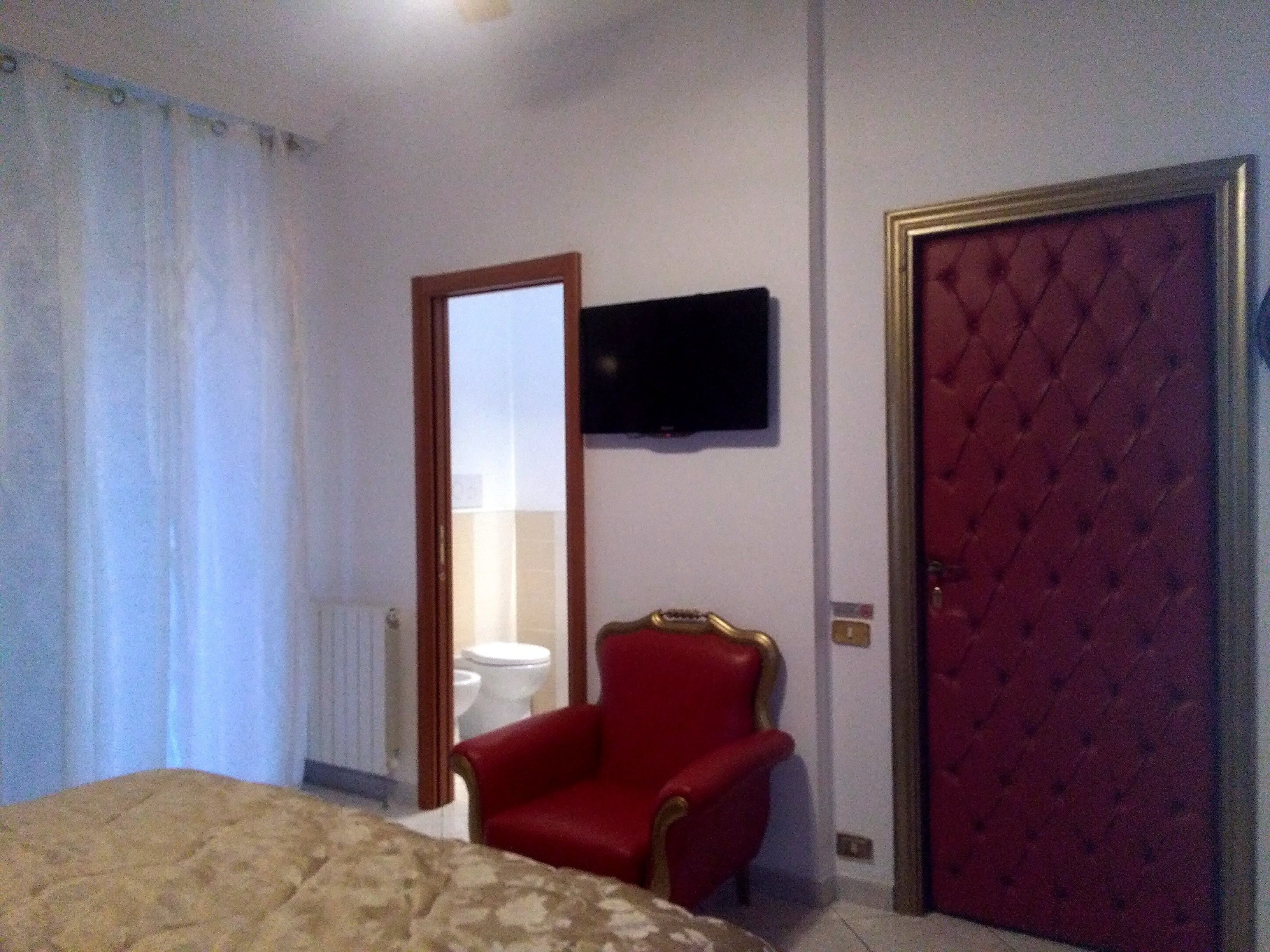 Una camera d'albergo con un letto, una poltrona rossa, un televisore a parete e una porta rossa.