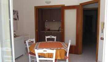 Un comedor con una mesa dispuesta para cuatro, una cocina al fondo y una puerta que da a otra habitación.