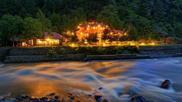 A riverside hotel with a prominent building illuminated against a forest backdrop. - Arcadian Riverside (Balakot)